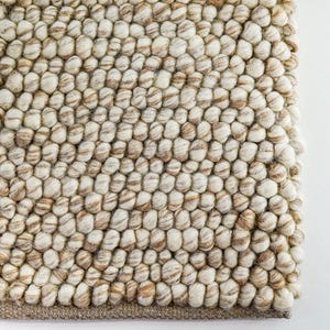 May include: A beige and brown wool rug with a textured, bumpy surface. The rug is made of small, round balls of wool that are woven together.