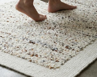 May include: A rectangular, off-white rug with a textured surface and a woven border. The rug features flecks of brown and tan throughout. The image shows a person's feet standing on the rug.