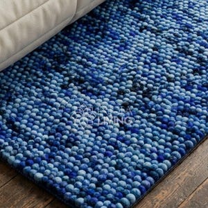 May include: A rectangular area rug composed of numerous small, spherical felted wool balls in varying shades of blue, ranging from light sky blue to deep navy. The rug features a white logo with the text INDIA LIVING printed in the center.