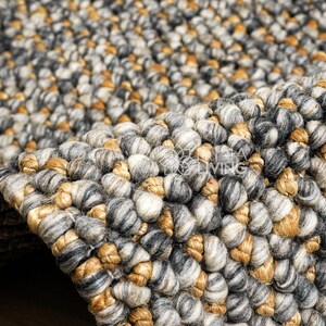 Puede incluir: Una alfombra gruesa tejida a mano con una construcci&oacute;n de pelo rizado grueso y texturizado. La alfombra presenta una paleta de colores variados en tonos gris carb&oacute;n, gris claro, blanco y amarillo ocre c&aacute;lido, creando una superficie densa y multidimensional.