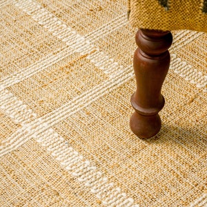 May include: A close-up of a natural fiber rug with a white woven pattern. The rug is beige and has a textured surface. A wooden chair leg is resting on the rug.