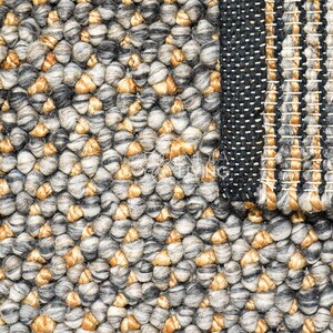 Puede incluir: Una alfombra texturizada con un tejido grueso de lana anudada a mano en una mezcla de tonos gris carb&oacute;n, gris claro y bronceado dorado. La superficie muestra un patr&oacute;n denso y granulado, con una secci&oacute;n que presenta un borde tejido a rayas.