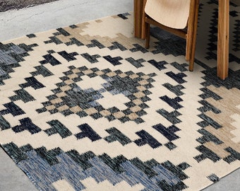 May include: A cream-coloured rug with a geometric pattern in shades of blue and brown. The rug is laid out on a concrete floor with a wooden chair and stool in the background.