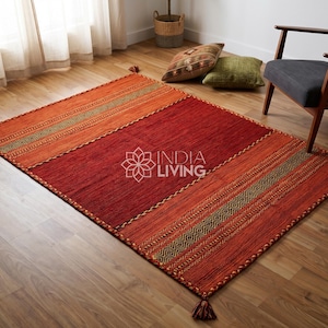 Op de afbeelding: Een rechthoekig vloerkleed met een groot centraal donkerrood paneel, geflankeerd door twee oranje secties met donkergroene geometrische patronen. Het kleed heeft kwastjes op elke hoek en toont de witte tekst INDIA LIVING in het midden.