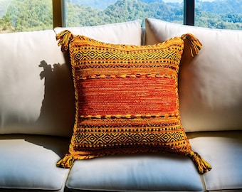 May include: A white couch with a colorful throw pillow with tassels. The pillow has a geometric pattern in shades of orange, yellow, and black. The couch is in front of a window with a view of a mountain range.