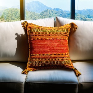 Puede incluir: Un sofá blanco con un cojín de colores con borlas. El cojín tiene un patrón geométrico en tonos naranja, amarillo y negro. El sofá está delante de una ventana con vistas a una cordillera.