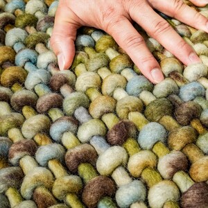 May include: Close-up of a hand resting on a woven rug with a textured, pebble-like design. The rug is made of wool in shades of green, brown, and blue.