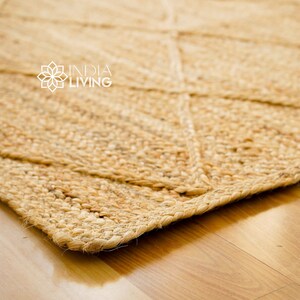May include: A close-up of a natural fiber rug with a diamond pattern. The rug is a light brown color and has a rough texture. The rug is made of jute and is perfect for adding a natural touch to any room. INDIA LIVING is printed in white text on the rug.