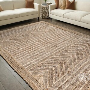 Può includere: Un tappeto rettangolare in juta con un motivo geometrico. Il tappeto presenta un colore beige naturale con un design intrecciato di linee diagonali e forme a spina di pesce. Il tappeto è posto su un pavimento in legno, con un divano sullo sfondo.