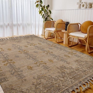 May include: A beige and gray area rug with a geometric pattern and fringe edges. The rug is laid out on a hardwood floor in a living room setting with two wicker chairs.