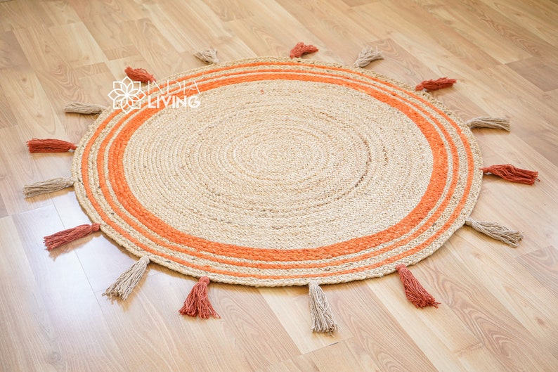 May include: A round jute rug with a natural beige color and orange stripes around the edge. The rug has tassels around the edge in both beige and orange.