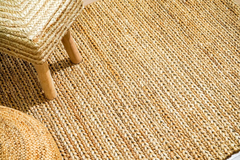 May include: A close-up shot of a natural fiber rug with a woven pattern. The rug is a light tan color and has a textured surface. A woven stool with wooden legs is partially visible in the upper left corner.