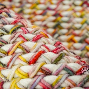 May include: A thick, hand-woven rug featuring a dense, braided texture. The weave consists of chunky white yarn intertwined with vibrant strands of red, yellow, green, and blue. The logo INDIA LIVING is visible in the bottom right corner.