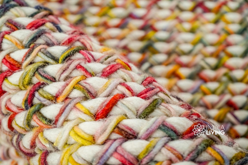 May include: A thick, hand-woven rug featuring a dense, braided texture. The weave consists of chunky white yarn intertwined with vibrant strands of red, yellow, green, and blue. The logo INDIA LIVING is visible in the bottom right corner.