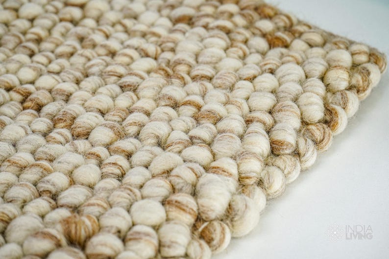 May include: A close-up of a beige and brown woven rug made of small, round, fluffy balls. The rug has a textured surface and a subtle pattern.