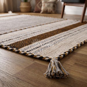 May include: A rectangular area rug featuring a woven pattern of cream, brown, and grey stripes with a braided border and corner tassels. The rug displays a repeating geometric texture and includes the text "KALA LIVING" printed in the center.