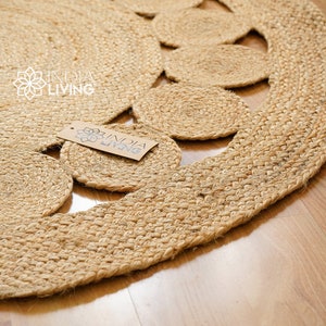May include: A round, natural jute rug with a decorative pattern of woven circles. The rug has a fringed edge and is made of natural fibers. The rug is labeled "India Living".