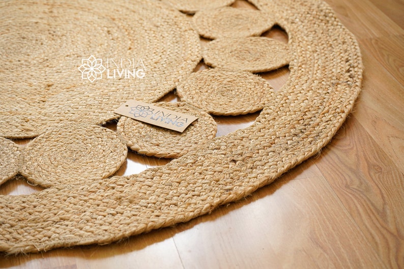 May include: A round, natural jute rug with a decorative pattern of woven circles. The rug has a fringed edge and is made of natural fibers. The rug is labeled "India Living".