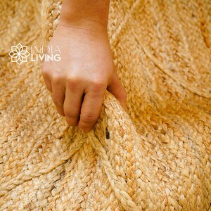 May include: Close-up of a natural jute rug with a hand holding the edge. The rug is woven with a natural, light brown color.