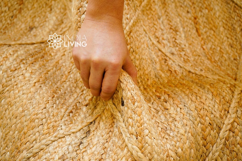 May include: Close-up of a natural jute rug with a hand holding the edge. The rug is woven with a natural, light brown color.