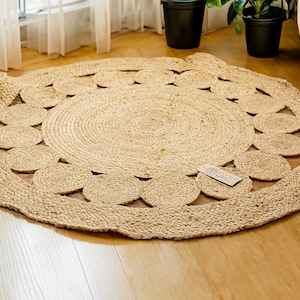 May include: A round, natural fiber rug with a woven design. The rug has a circular pattern of cutouts around the edge, creating a decorative border. The rug is made of natural jute fibers and has a light brown color.