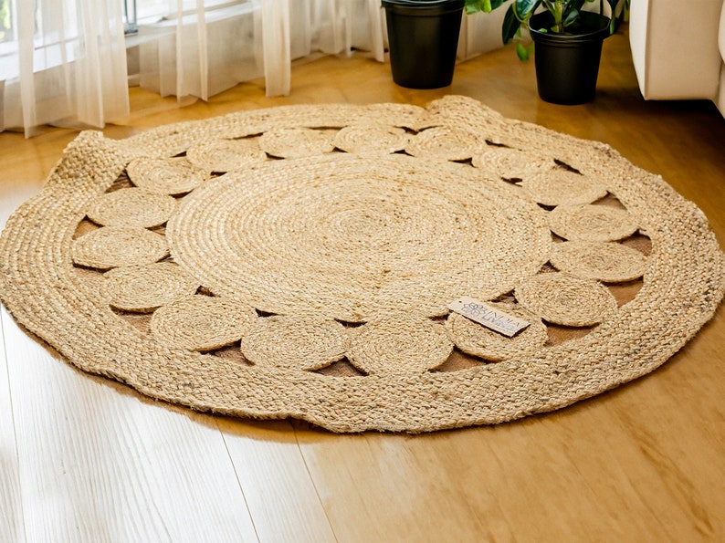 May include: A round, natural fiber rug with a woven design. The rug has a circular pattern of cutouts around the edge, creating a decorative border. The rug is made of natural jute fibers and has a light brown color.