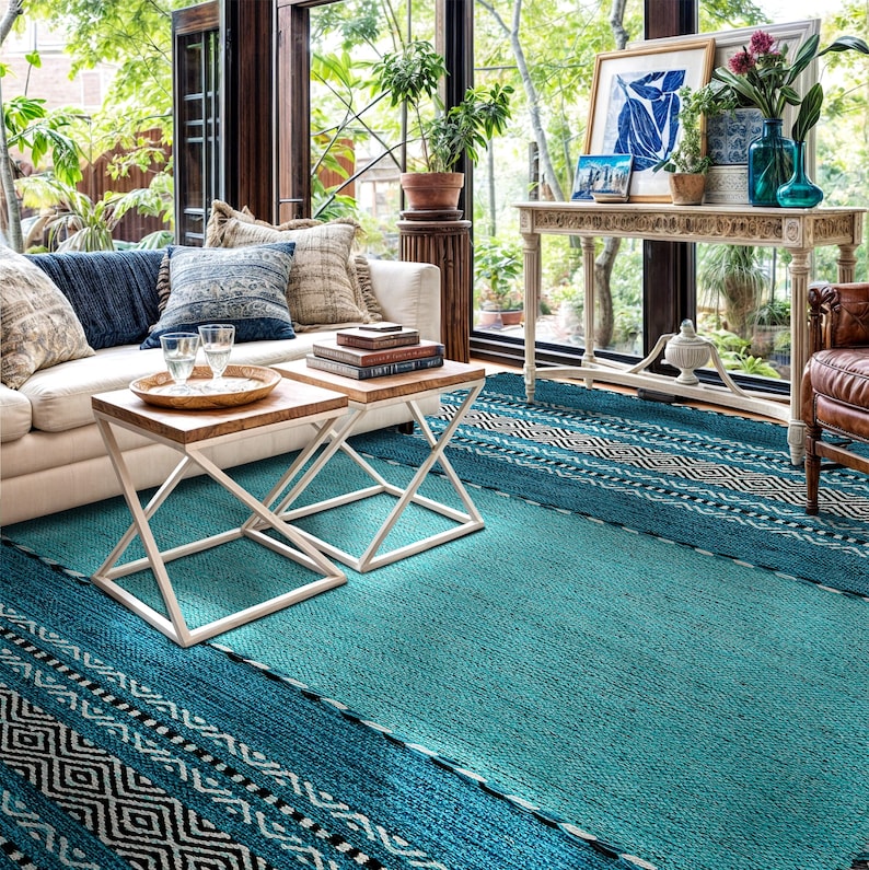 May include: A large rectangular area rug featuring a vibrant teal center with textured weave, bordered by wide bands of black and white geometric diamond patterns. The rug covers the floor space, showcasing a distinct contrast between the solid teal field and the intricate, repeating tribal-inspired motifs along the edges, creating a bold visual anchor for the room.