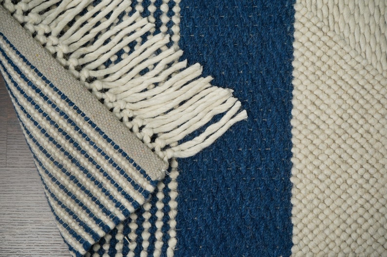 May include: A white and blue striped rug with a fringe border. The rug has a textured weave and is made of natural fibers.