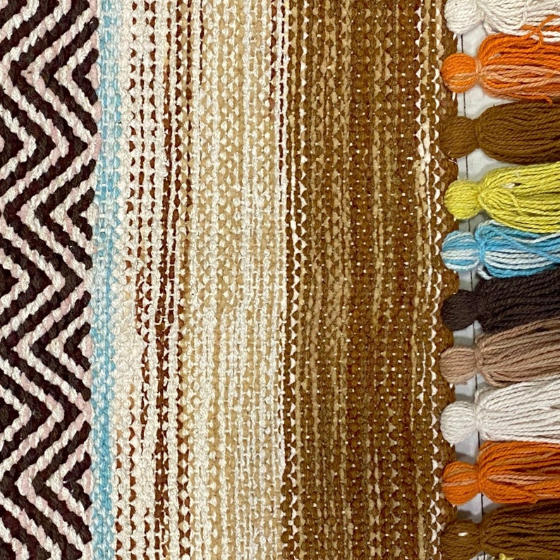 May include: A woven rug with a brown, beige, and blue striped pattern. The rug has a fringe with tassels in various colors, including orange, yellow, blue, brown, and white.