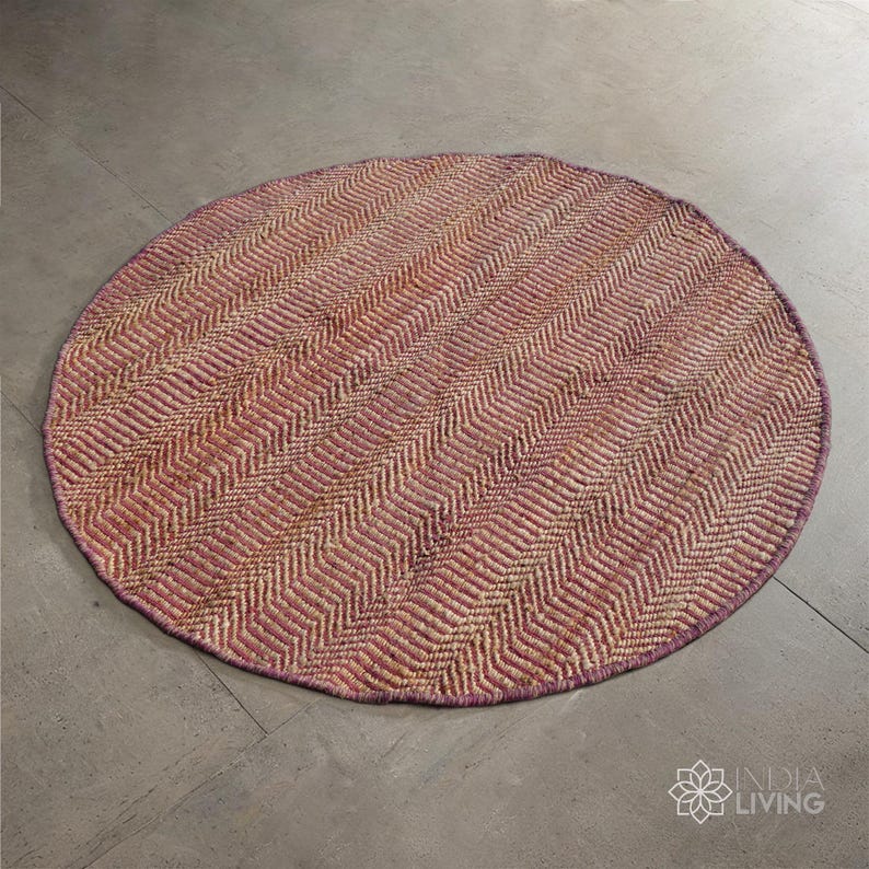 May include: A round, woven rug with a herringbone pattern in shades of pink and beige. The rug has a fringed edge.