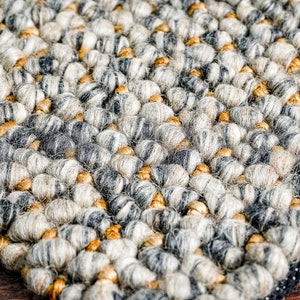 Puede incluir: Una alfombra gruesa y texturizada con un patr&oacute;n denso de fibras redondeadas y anudadas en tonos gris carb&oacute;n, gris claro y crema, intercaladas con peque&ntilde;os acentos suaves de color marr&oacute;n dorado en toda la superficie tejida.