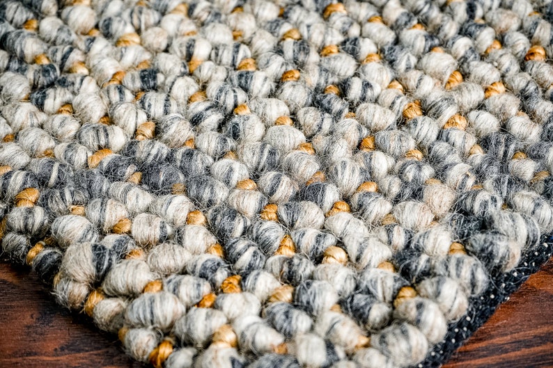 Puede incluir: Una alfombra gruesa y texturizada con un patr&oacute;n denso de fibras redondeadas y anudadas en tonos gris carb&oacute;n, gris claro y crema, intercaladas con peque&ntilde;os acentos suaves de color marr&oacute;n dorado en toda la superficie tejida.