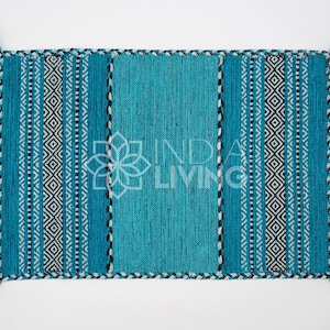 May include: A rectangular woven rug in shades of teal and turquoise, featuring a central solid panel flanked by vertical stripes and geometric diamond patterns in black and white. The edges are finished with a braided border and teal tassels.
