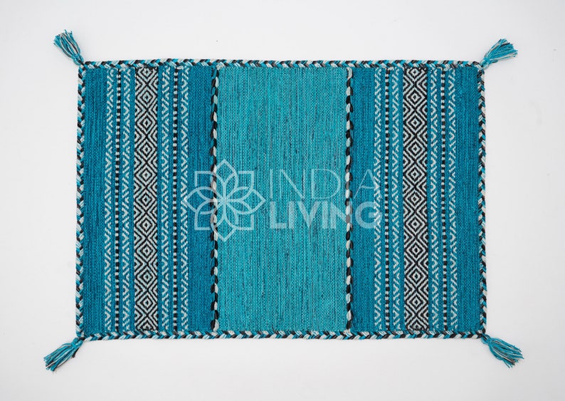 May include: A rectangular woven rug in shades of teal and turquoise, featuring a central solid panel flanked by vertical stripes and geometric diamond patterns in black and white. The edges are finished with a braided border and teal tassels.