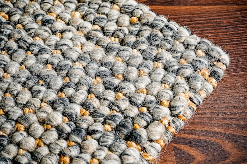 Puede incluir: Una alfombra texturizada con una construcci&oacute;n densa anudada a mano de fibras de lana gruesas y redondeadas. La superficie muestra una paleta de colores variados en gris carb&oacute;n, gris claro, crema y tonos bronce c&aacute;lidos en un patr&oacute;n.