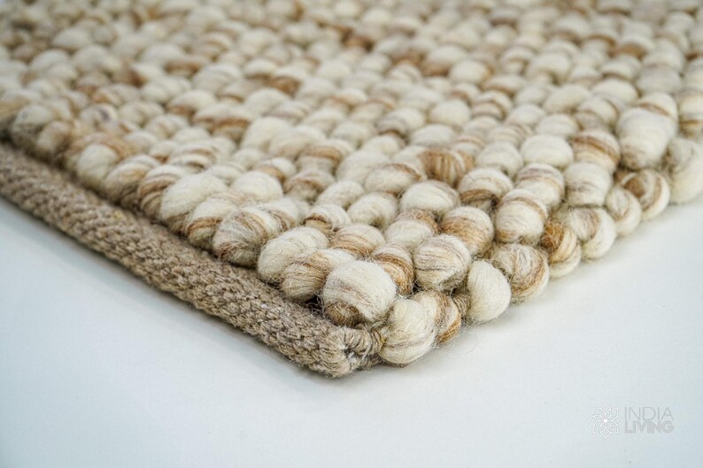 K&ouml;nnte beinhalten: Nahaufnahme eines beige-wei&szlig;en Woll-Teppichs mit einer strukturierten, geknoteten Oberfl&auml;che. Der Teppich hat einen braunen gewebten Rand.