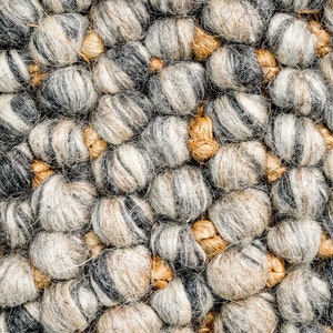 Puede incluir: Una alfombra texturizada compuesta por bolas de lana afieltrada esf&eacute;ricas y densas en tonos gris carb&oacute;n, gris claro y blanco, intercaladas con nudos de yute o fibra m&aacute;s peque&ntilde;os, lisos y de color tostado que conectan los elementos de lana.