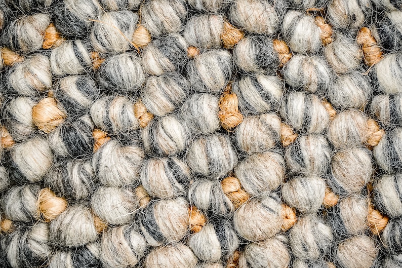 Puede incluir: Una alfombra texturizada compuesta por bolas de lana afieltrada esf&eacute;ricas y densas en tonos gris carb&oacute;n, gris claro y blanco, intercaladas con nudos de yute o fibra m&aacute;s peque&ntilde;os, lisos y de color tostado que conectan los elementos de lana.