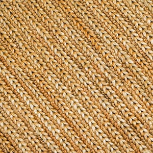 May include: Close-up of a woven material with a diagonal pattern. The texture is made of thick, light brown fibers, creating a natural, rustic appearance. The weave is tight and consistent, suggesting a durable, handcrafted item, possibly a rug or placemat.