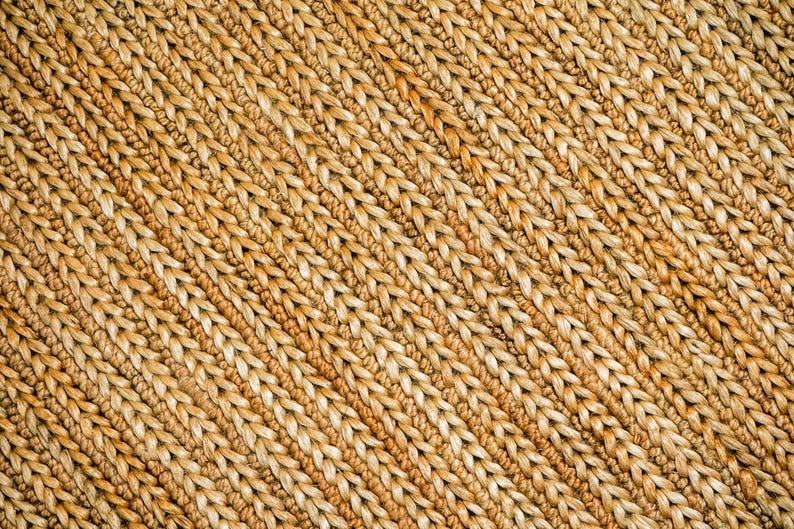 May include: Close-up of a woven material with a diagonal pattern. The texture is made of thick, light brown fibers, creating a natural, rustic appearance. The weave is tight and consistent, suggesting a durable, handcrafted item, possibly a rug or placemat.