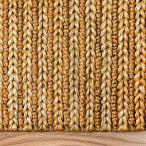 May include: Close-up of a woven rug with a textured surface. The rug is made of natural fibers in shades of tan and gold, with a repeating pattern of vertical braids. The rug is placed on a wooden floor.