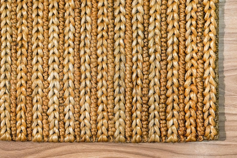 May include: Close-up of a woven rug with a textured surface. The rug is made of natural fibers in shades of tan and gold, with a repeating pattern of vertical braids. The rug is placed on a wooden floor.