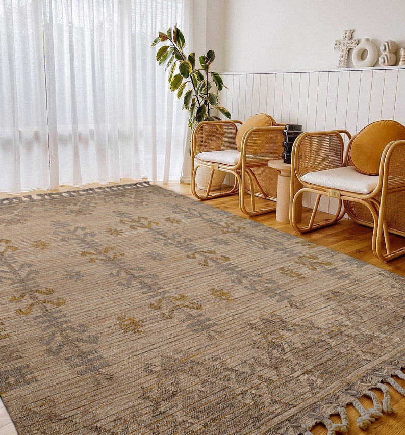 May include: A beige and gray area rug with a geometric pattern and fringe edges. The rug is laid out on a hardwood floor in a living room setting with two wicker chairs.