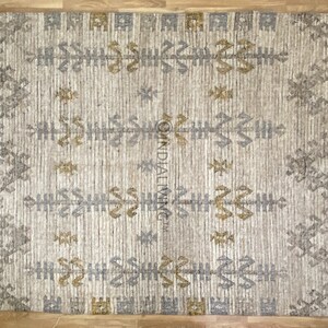 May include: A beige rug with a geometric pattern in shades of gray and gold. The rug has fringe on all sides.