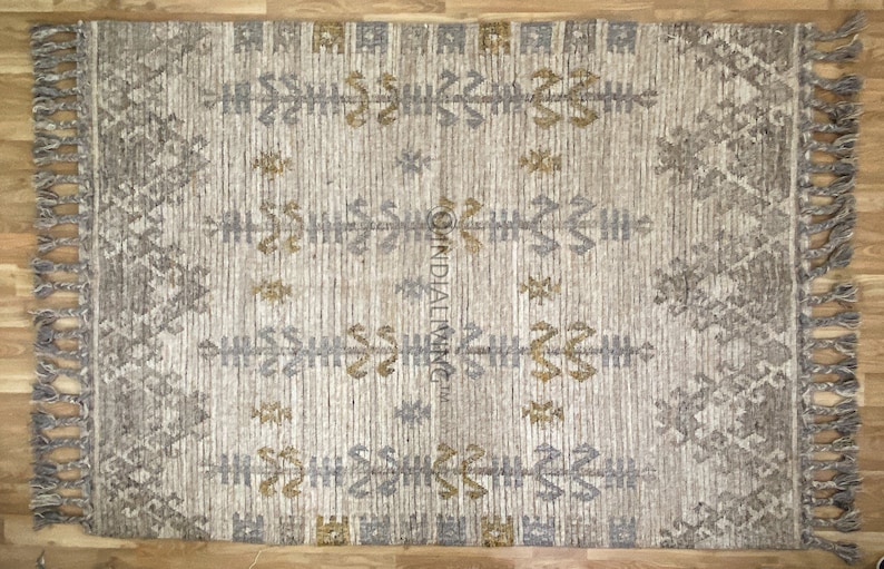 May include: A beige rug with a geometric pattern in shades of gray and gold. The rug has fringe on all sides.