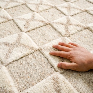 May include: A close-up of a beige and white woven rug with a geometric pattern. The rug has a soft, textured surface.
