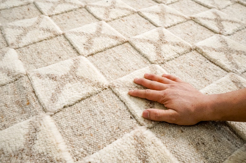 May include: A close-up of a beige and white woven rug with a geometric pattern. The rug has a soft, textured surface.