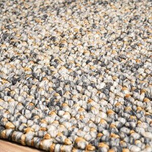 Puede incluir: Una alfombra gruesa tejida a mano con un patr&oacute;n de punto grueso y texturizado. La alfombra est&aacute; compuesta por una densa mezcla de hilos en tonos gris carb&oacute;n, gris claro, crema y amarillo mostaza c&aacute;lido, creando un aspecto variado y tonal.