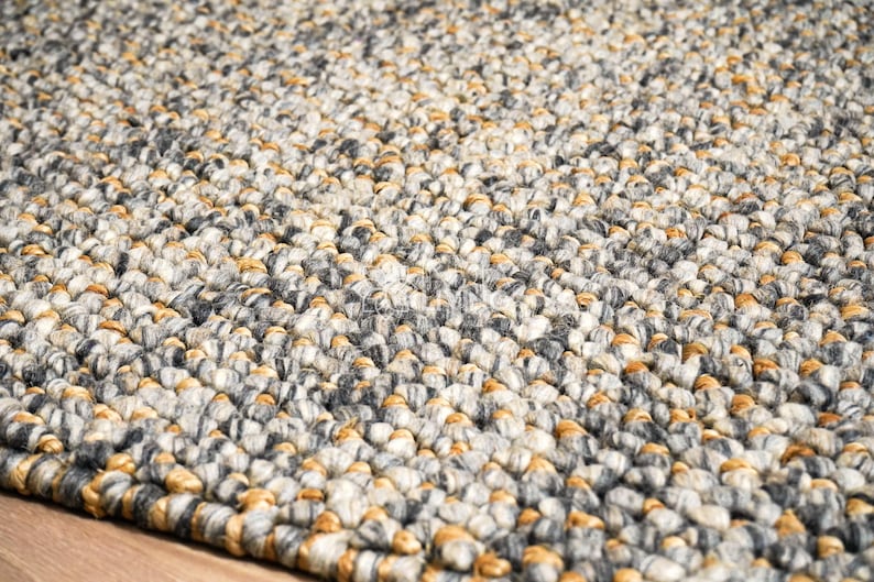 Puede incluir: Una alfombra gruesa tejida a mano con un patr&oacute;n de punto grueso y texturizado. La alfombra est&aacute; compuesta por una densa mezcla de hilos en tonos gris carb&oacute;n, gris claro, crema y amarillo mostaza c&aacute;lido, creando un aspecto variado y tonal.