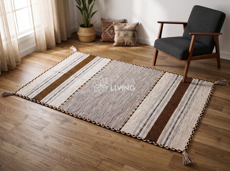 May include: A rectangular area rug featuring a central gray section flanked by cream and brown striped panels with geometric patterns. The rug has a braided border and tassels at each corner, with the text INDIA LIVING printed in the center.